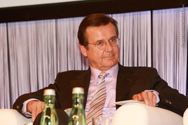 Ein Mann mit Brille und Anzug sitzt auf einem weißen Sessel.