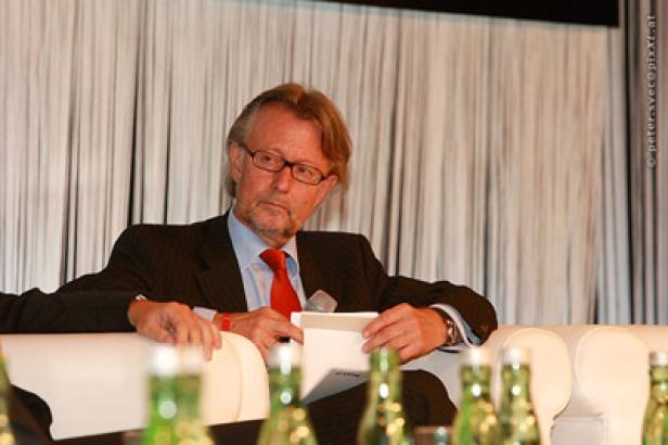 Ein Mann mit Brille und Anzug sitzt auf einem Sofa und hält ein Dokument in der Hand.