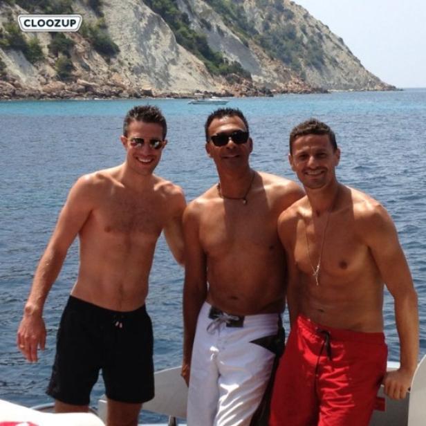 Drei Männer stehen barfuß auf einem Boot vor einer Küstenlandschaft.