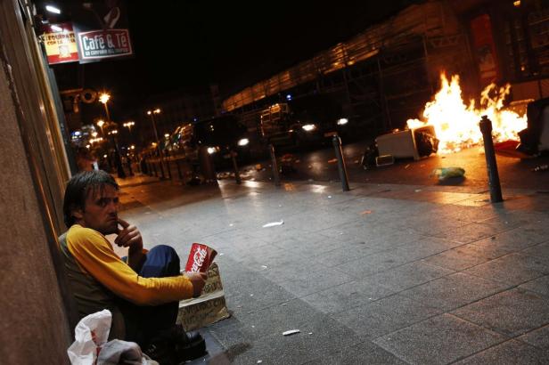 Ein Mann sitzt nachts auf einer Straße, im Hintergrund brennt ein Feuer.