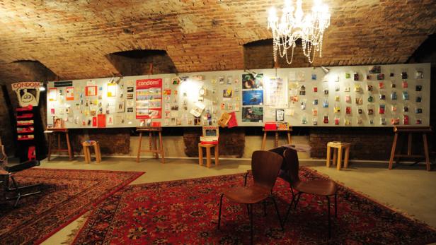 Ein Ausstellungsraum mit Kondomverpackungen an einer Wand, Teppichen und einem Kronleuchter.