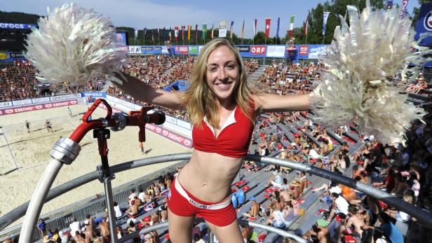 Eine Cheerleaderin mit Pompons steht vor einem Publikum bei einem Beachvolleyballspiel.