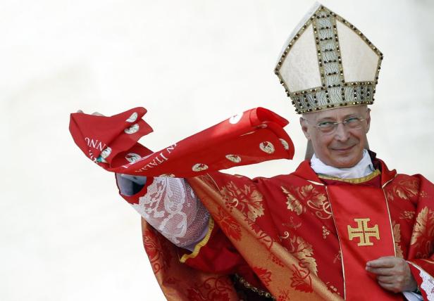 Ein Bischof in roter Robe und Mitra hält ein rotes Tuch mit der Aufschrift „In Italia“.