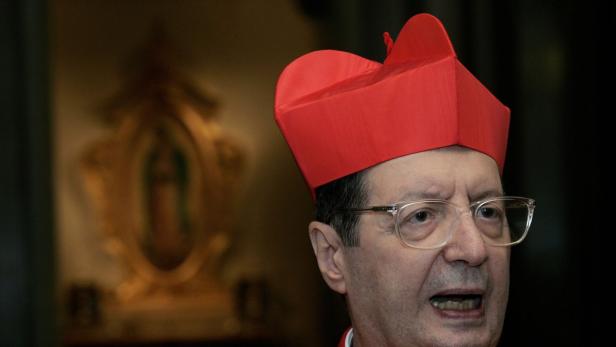 Ein Kardinal mit Brille und rotem Birett vor einem goldenen religiösen Ornament.