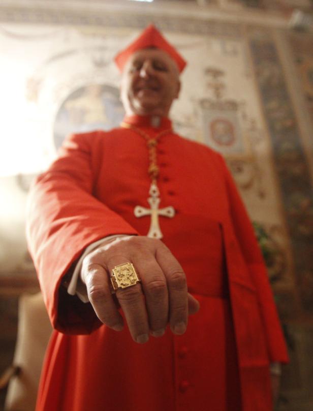 Ein Kardinal in roter Kleidung zeigt seinen goldenen Ring.