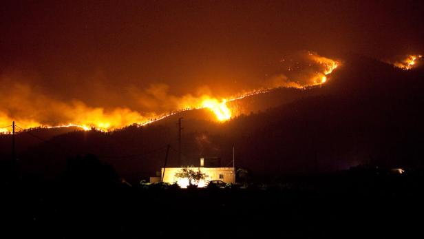 Ein Waldbrand erstreckt sich nachts über einen Hügel, im Vordergrund ein beleuchtetes Haus.