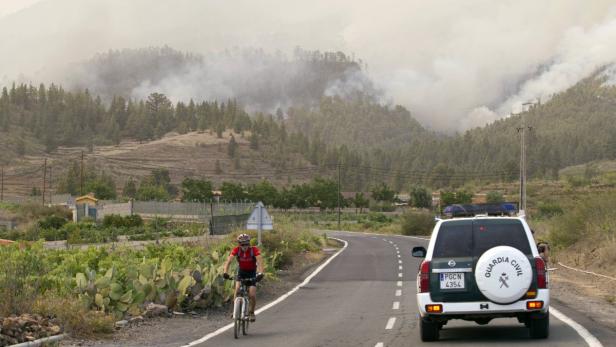 Ein Radfahrer fährt an einem Auto der Guardia Civil vorbei, während im Hintergrund ein Waldbrand zu sehen ist.