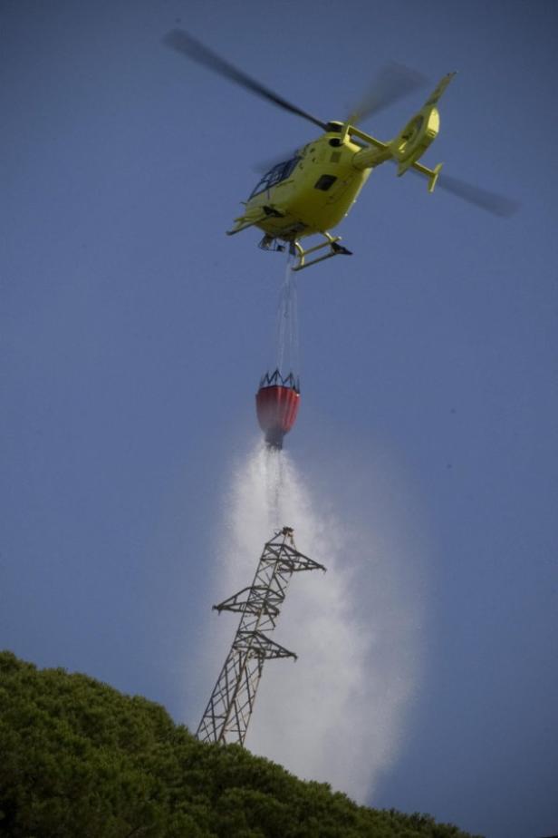 Ein gelber Hubschrauber löscht mit einem Wasserbehälter einen Brand an einem Strommast.