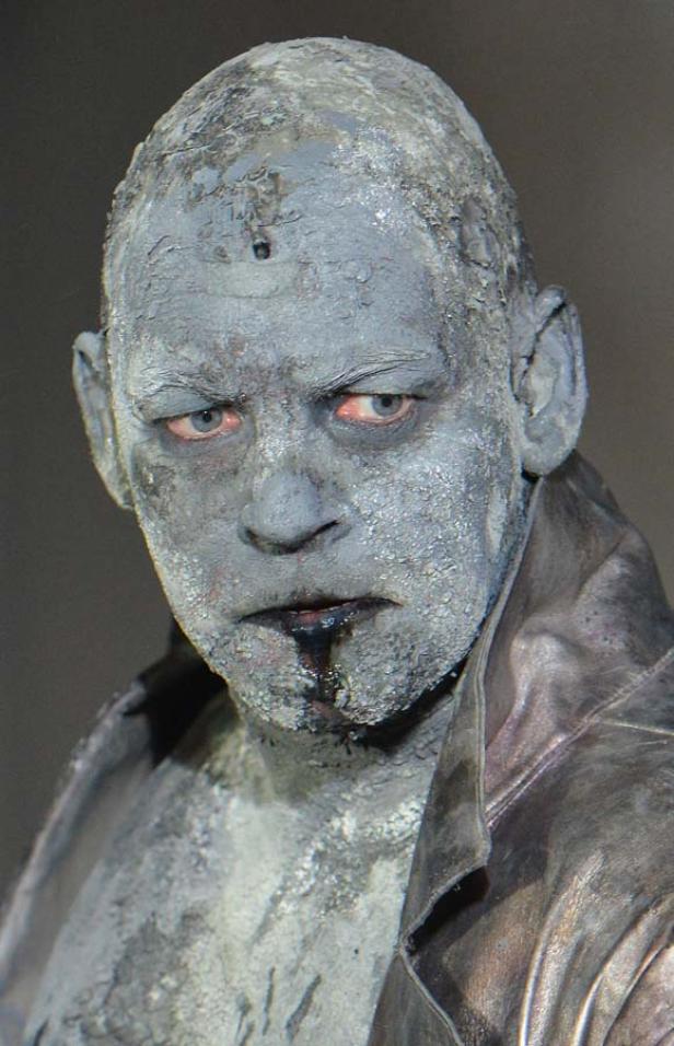 Ein Mann mit grauer Farbe im Gesicht und blutigem Mund blickt grimmig.