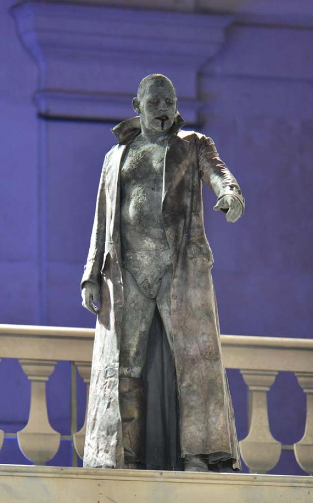 Ein als Statue geschminkter Mann in Unterwäsche und Mantel steht auf einem Podest.
