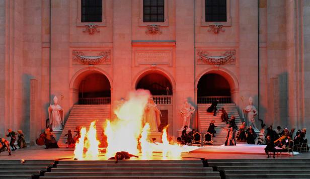 Eine Theaterszene mit Schauspielern, Statuen und Feuer vor einem großen Gebäude.
