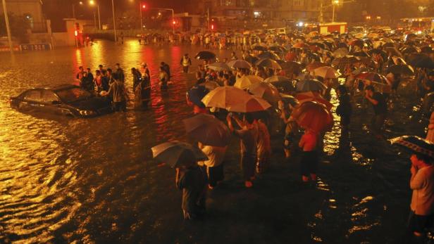 Eine überflutete Straße bei Nacht mit Menschen, die mit Regenschirmen durch das Wasser waten, und einem teilweise versunkenen Auto.