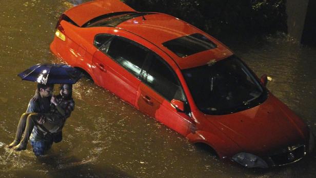 Ein Mann trägt eine Frau durch das Hochwasser, neben einem roten Auto.