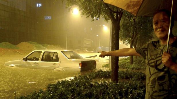 Ein Mann steht im Regen mit einem Regenschirm, während ein Auto im Hochwasser steht.