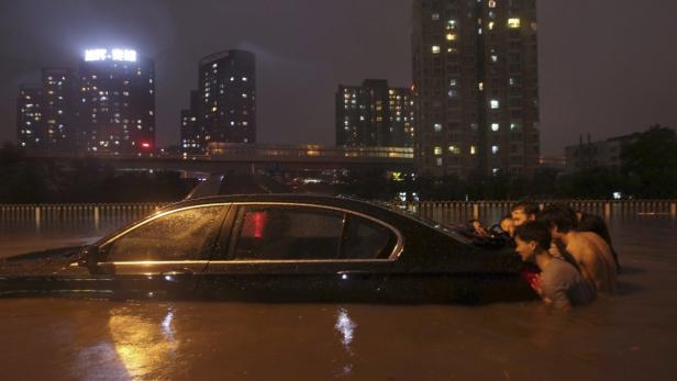 Ein Auto ist bis zum Dach in einem städtischen Gebiet überflutet, während Menschen im Wasser stehen.