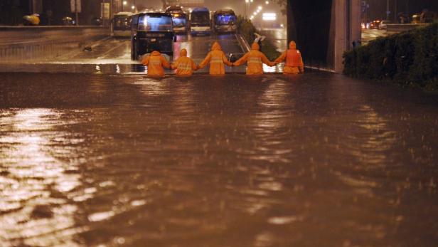 Fünf Personen in Regenkleidung stehen Händchen haltend in einer überfluteten Straße.