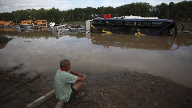 Ein Mann sitzt am Ufer eines überfluteten Gebiets, in dem Autos und ein Bus im Wasser stehen.