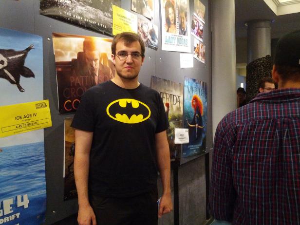 Ein Mann mit einem Batman-T-Shirt steht vor einer Wand mit Filmplakaten, darunter „Merida“ und „Ice Age 4“.