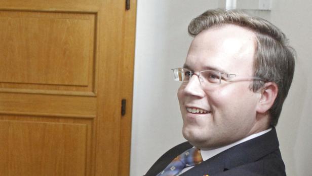 Ein lächelnder Mann mit Brille und Anzug vor einer Holztür.