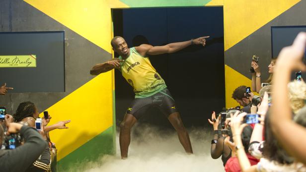 Usain Bolt posiert vor einer Menschenmenge mit Handykameras vor einem jamaikanisch gestreiften Hintergrund.