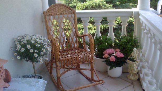 Ein Schaukelstuhl aus Rattan steht auf einem Balkon, umgeben von Blumen und Dekorationen.