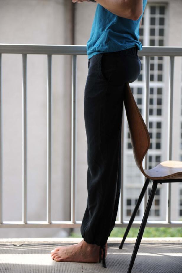 Eine Person steht barfuß auf einem Balkon, ein Stuhl ist zwischen ihren Beinen eingeklemmt.