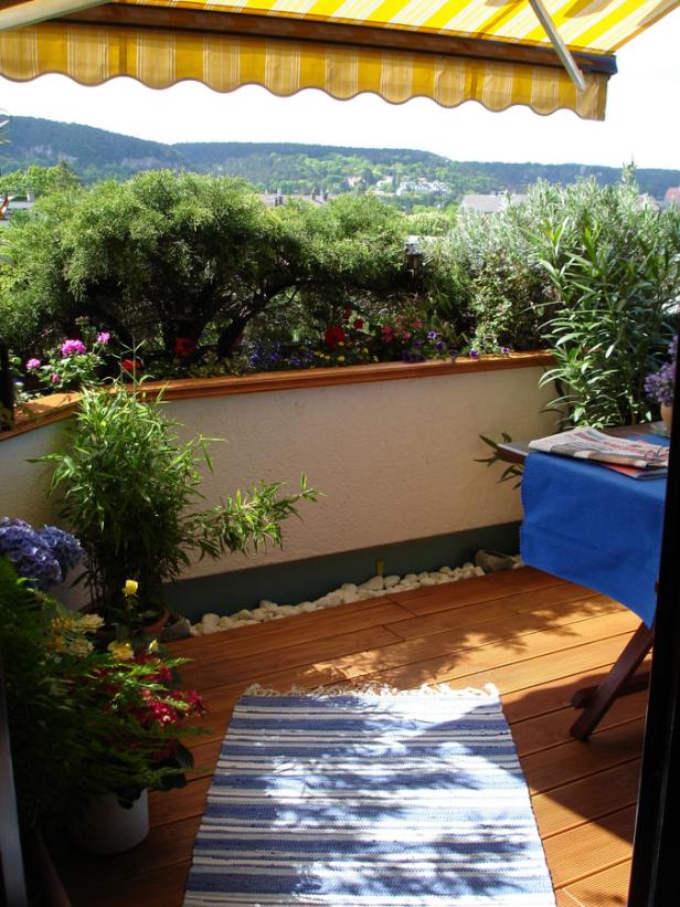 Ein sonniger Balkon mit Holzboden, Pflanzen und Blick auf eine grüne Hügellandschaft.