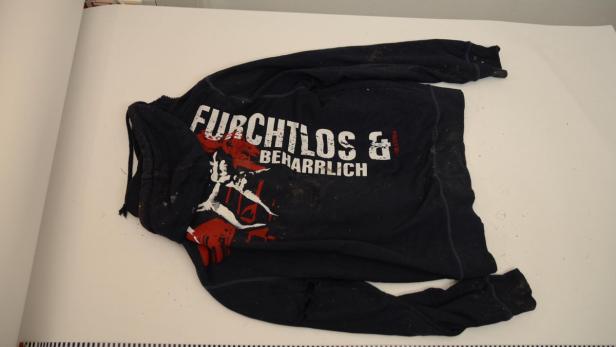 Ein dunkler, verschmutzter Pullover mit der Aufschrift „Furchtlos & Beharrlich“ liegt auf einem Tisch.