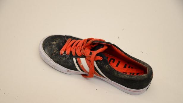 Ein schmutziger, schwarzer Turnschuh mit orangefarbenen Schnürsenkeln liegt auf einem hellen Untergrund.