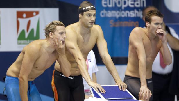 Drei Schwimmer stehen an den Startblöcken vor einem Wettkampf.