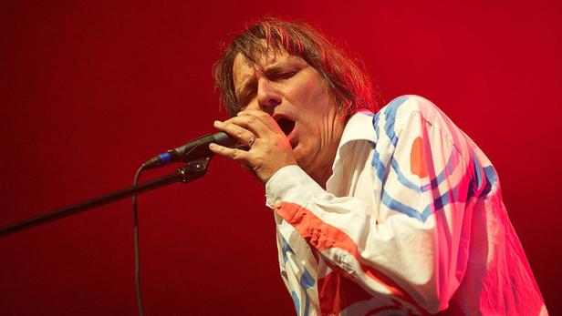 Ein Sänger mit offenem Mund steht vor einem Mikrofon auf einer Bühne.