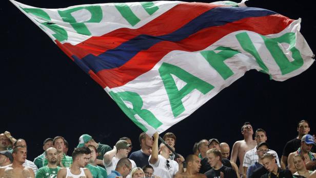 Eine Gruppe von Fußballfans schwenkt eine große Flagge mit den Farben Rot, Weiß und Blau sowie dem Schriftzug „RAPID“.