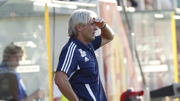 Ein Trainer mit grauen Haaren schaut mit der Hand an der Stirn auf das Spielfeld.