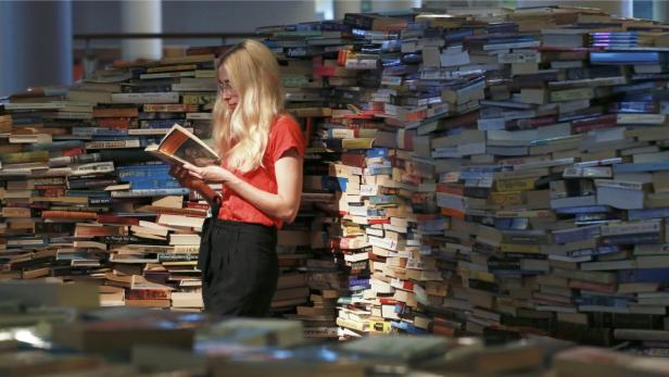 Eine Frau mit blonden Haaren liest ein Buch vor einem riesigen Stapel Bücher.