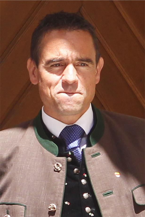 Ein Mann in Tracht mit Krawatte vor einer Holzwand.