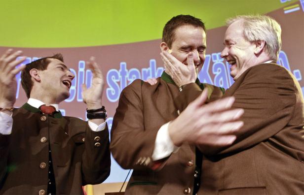 Drei Männer in Trachtenjacken lachen und gestikulieren vor einem Bildschirm.