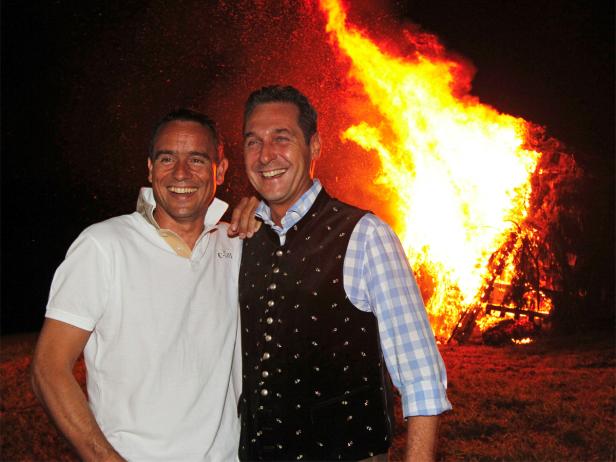 Zwei Männer lächeln vor einem großen Lagerfeuer.