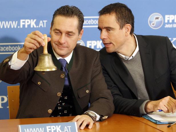 Zwei Männer sitzen an einem Tisch, einer läutet eine Glocke.