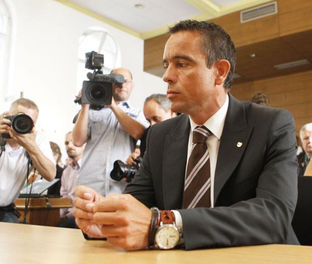 Ein Mann im Anzug sitzt an einem Tisch, umringt von Fotografen.