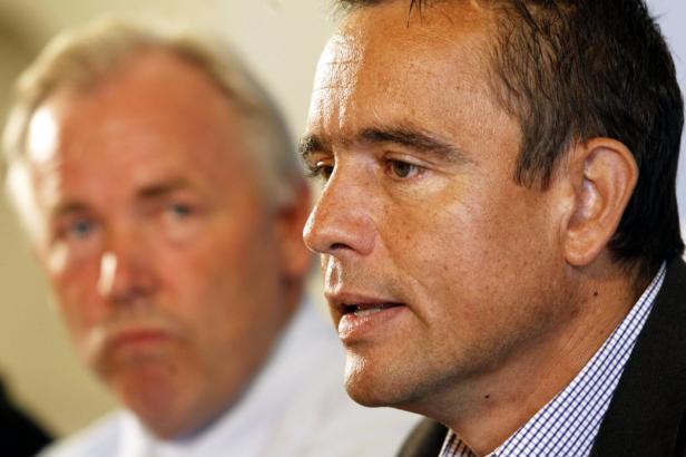 Ein Mann spricht, während eine andere Person im Hintergrund unscharf ist.