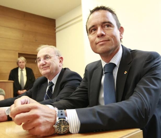 Zwei Männer in Anzügen sitzen an einem Tisch, ein dritter Mann steht im Hintergrund.