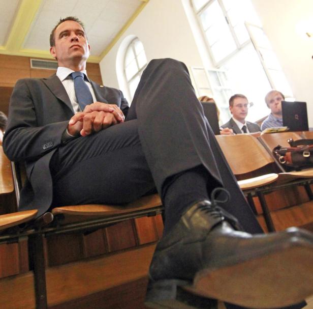 Ein Mann im Anzug sitzt mit verschränkten Händen in einem Raum voller Menschen.