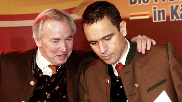 Zwei Männer in Trachtenjacken stehen nebeneinander vor einem Wahlplakat.