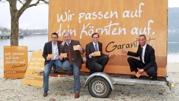 Vier Männer posieren vor einem Anhänger mit der Aufschrift „Wir passen auf dein Kärnten auf“.