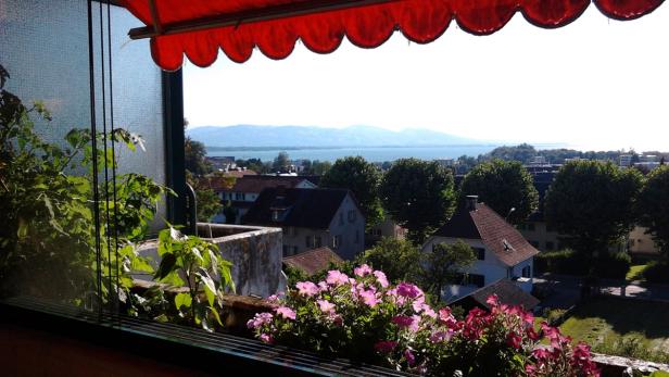 Blick von einem Balkon mit Blumen auf eine Stadt am See mit Bergen im Hintergrund.