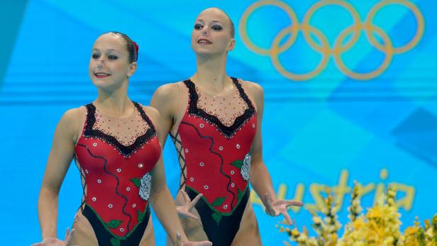 Zwei Synchronschwimmerinnen in roten Badeanzügen mit Blumenmuster vor blauem Hintergrund.