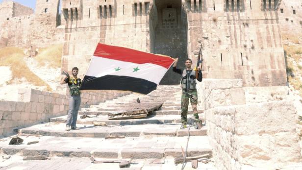 Zwei bewaffnete Männer halten eine syrische Flagge vor einer Burg.