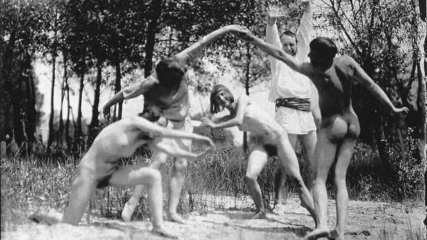 Eine Gruppe nackter Menschen tanzt im Wald, eine Person ist bekleidet.