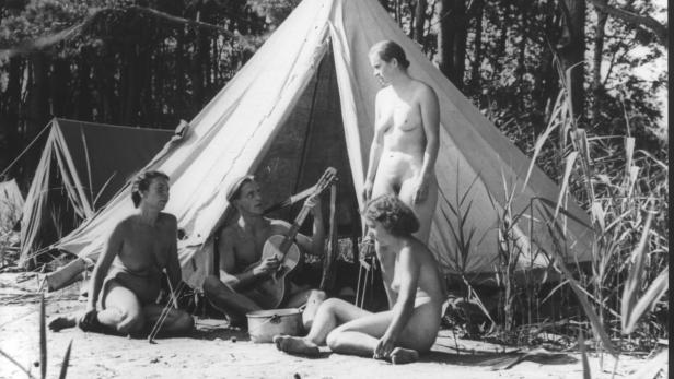 Eine Gruppe von Menschen campt nackt an einem See, ein Mann spielt Gitarre.