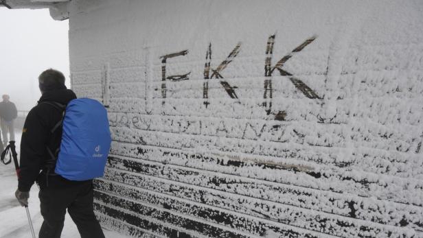 Ein Wanderer mit Rucksack steht vor einer schneebedeckten Holzwand mit der Aufschrift „EKK“.